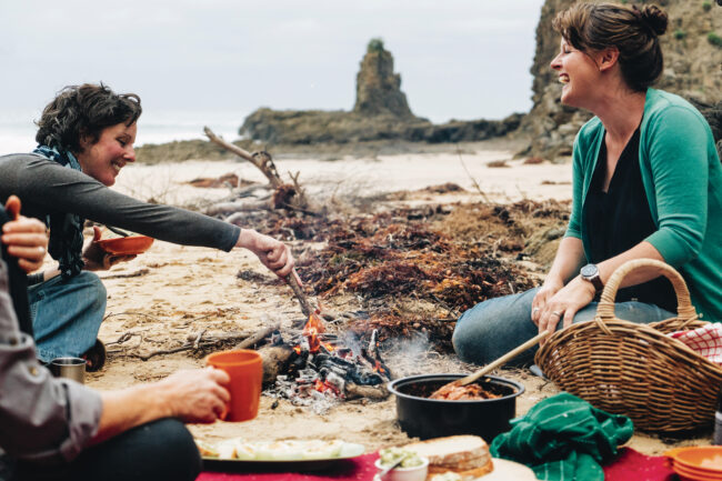 Kirsten Bradley & Fiona Weir sharing a joyful meal by a campfire at Kiama beach. Image from a post on sustainable holiday projects.