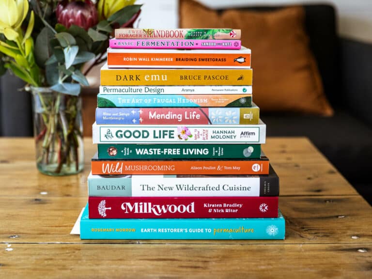 A tall stack of gardening and permaculture books on a table, including Milkwood and Hannah Moloney's The Good Life