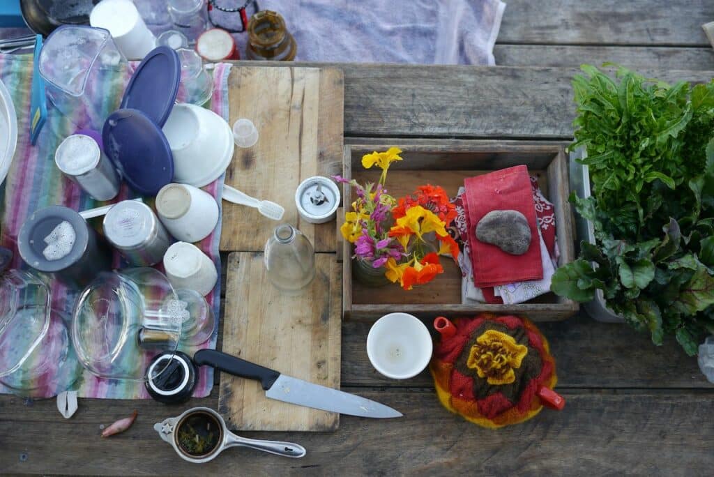 Annie & Genevieve's wash up. An outdoor kitchen scene with various cups, crockery & utensils spread on tea towels on a table. A pot of tea and salad greens.