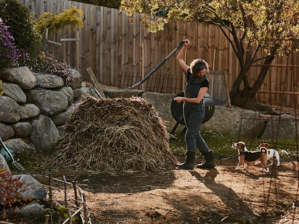 How to compost dog poop - a woman i overalls with a garden fork and compost pile, looks at two small dogs