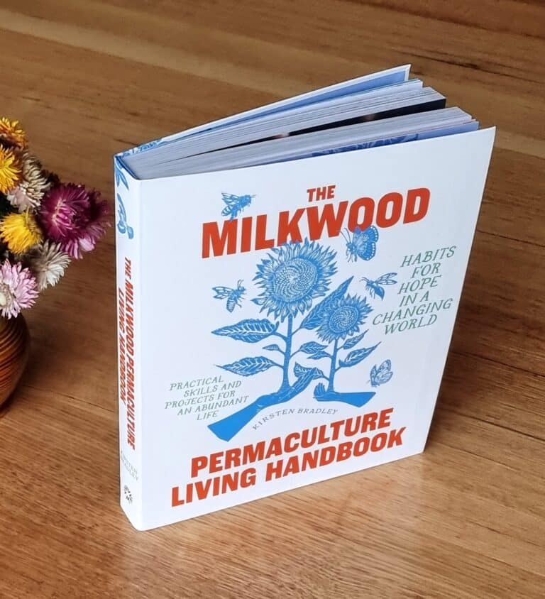 The Permaculture Living Handbook standing on a wooden table beside a vase of dried flowers